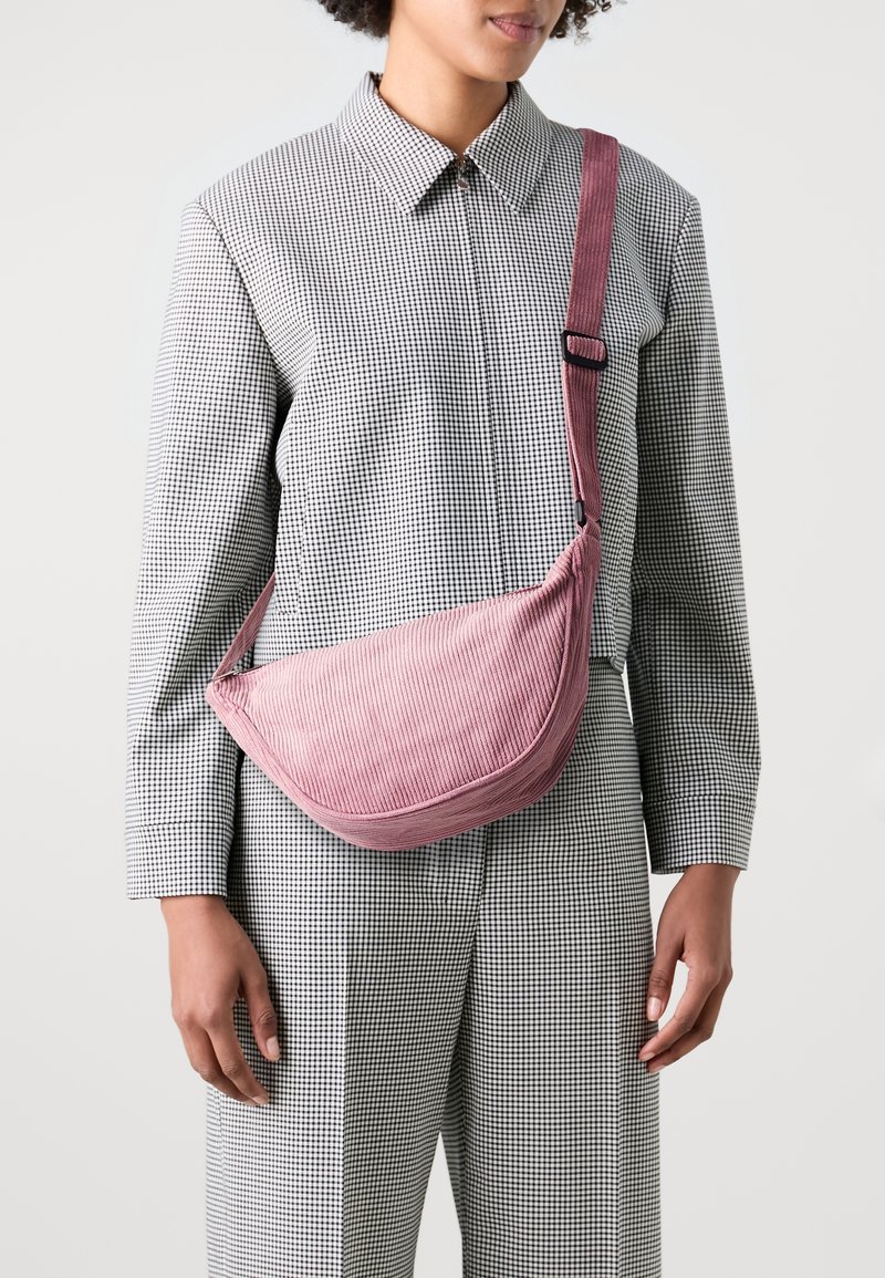 Kompaktní růžová manšestrová kabelka přes rameno s zakřiveným tvarem, nastavitelným popruhem a texturovaným povrchem, nošená přes černo-bílé kostkované oblečení.