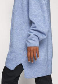 Ljusblå stickad tröja med långa ärmar, ribbade manschetter och sidospel. Mjuk textur, avslappnad passform och något förlängd bakhel.