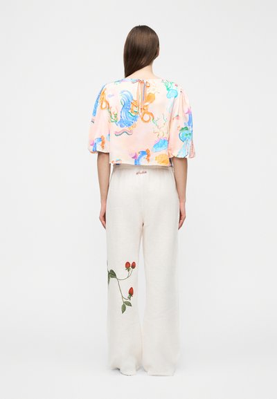 Femme aux longs cheveux bruns portant un haut pastel avec un motif de vie marine et un pantalon blanc à jambes larges avec une broderie de baies rouges, vue de dos.