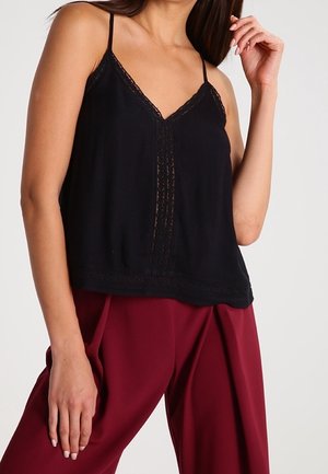 Vrouw draagt een zwart mouwloos topje met kanten rand, gecombineerd met hoog getailleerde wijde bordeauxrode broek.