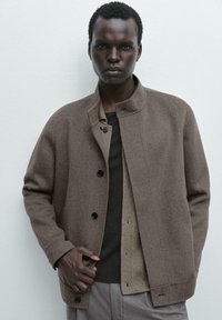 Brown wool-blend jacket with a high collar and black buttons, layered over a dark knit shirt and light textured layer underneath.