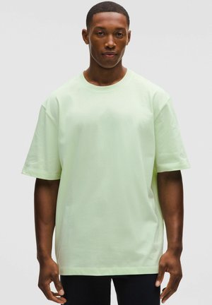 Mand iført en ensfarvet lys grøn oversized t-shirt med korte ærmer, stående mod en neutral grå baggrund.