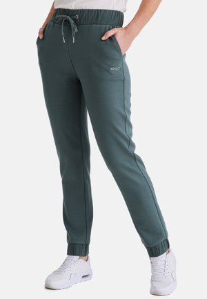 Person trägt dunkelgrüne Joggerhosen mit elastischen Bündchen, Kordelzug in der Taille, Seitentaschen und weißen Sportschuhen.