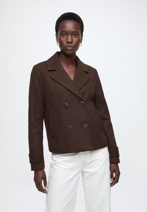 Femme portant une veste croisée marron foncé avec de gros boutons et un pantalon blanc sur un fond clair uni.