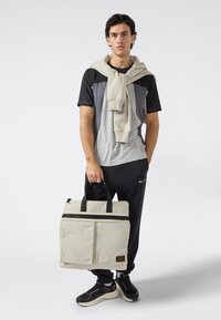 Joven en ropa deportiva casual sosteniendo una bolsa de lona beige, con un sudadera beige colgada sobre sus hombros, contra un fondo blanco.