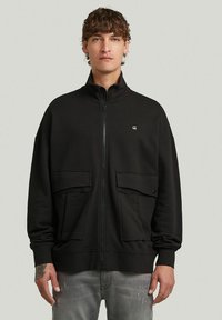 Sweat-shirt zippé noir avec un col montant, comportant deux poches avant et un petit logo sur la poitrine. Matière douce et texturée.