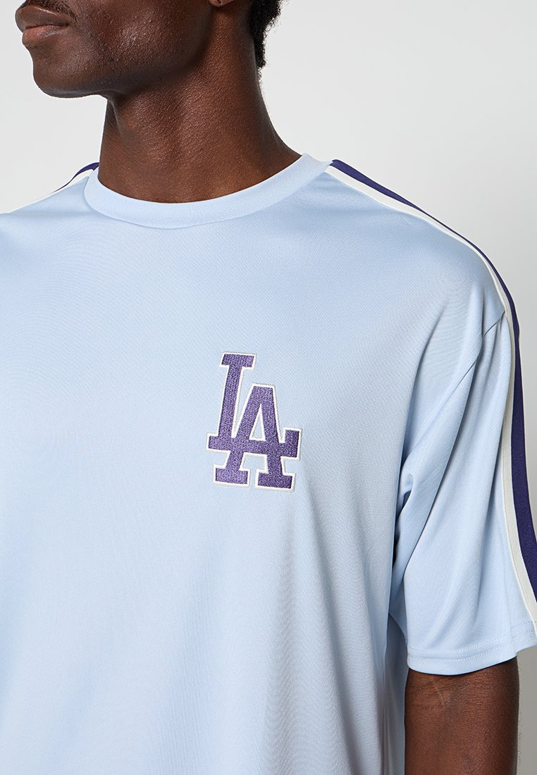 Jasnoniebieska koszulka sportowa wykonana z oddychającego materiału, z fioletowym logo "LA" i kontrastującymi ciemnoniebieskimi paskami po bokach.