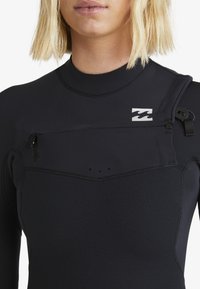 Nahaufnahme einer Person in einem schwarzen Neoprenanzug mit Reißverschluss und Logo auf der Brust, kurzes blondes Haar vor weißem Hintergrund sichtbar.