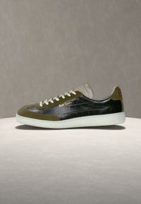 Baskets basses avec motif crocodile noir, détails en daim olive, semelle blanche et lacets, et logo doré "Paul Smith" sur fond neutre.