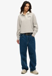 Sudadera tipo polo corta de manga larga en gris claro con un color blanco y el logo de "Superdry"; combinada con jeans azules holgados y zapatillas beige.