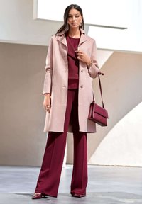Heller rosa Wollmantel mit Kragen, getragen über einem tief burgunderfarbenen Oberteil und weit geschnittenen Hosen. Accessoires aus einer passenden Handtasche und spitzen Schuhen.