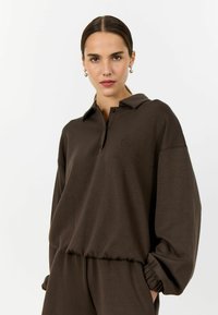 Hnědý polo top s dlouhým rukávem a límečkem, zapínáním na knoflíky a žebrovanými manžetami. Obsahuje jemný detail loga na hrudi. Měkká textilie.
