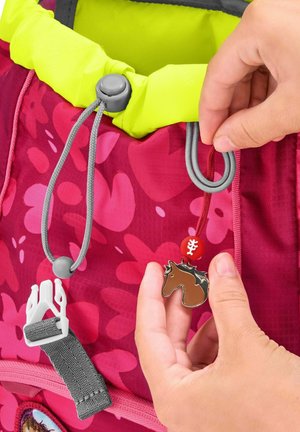 Rosa Rucksack mit floralem Muster, gelbem Oberteil, grauer Hardware und einem braunen, pferdeförmigen Anhänger, der in der Nähe einer dekorativen Schlaufe gehalten wird.