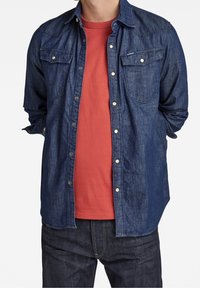 Tumman denim-kankaan nappikauluspaita, jossa on kaksi rintataskua, kerroksittain punaisen yksivärisen t-paidan päällä. Sisältää klassisen kauluksen ja nappikiinnityksen.