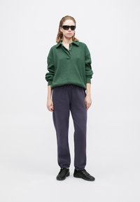 Suéter de textura verde con cuello y dos botones, combinado con pantalones de chándal gris oscuro y zapatillas negras. Gafas de sol negras como acento.