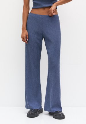 Vrouw die blauwe, geribbelde wijde pijpen broek en zwarte stevige schoenen draagt, staand tegen een effen witte achtergrond, hand rustend nabij de tailleband.