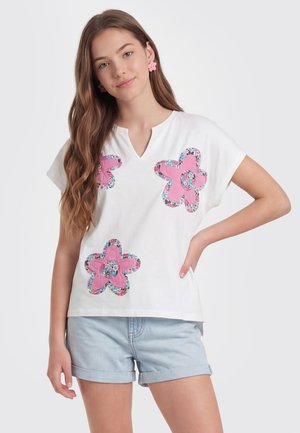 Jeune femme aux longs cheveux portant une chemise blanche avec des patchs floraux roses et un short en jean bleu clair, debout avec une main sur la hanche.