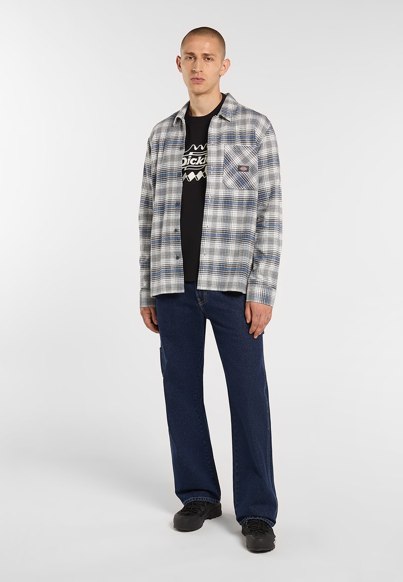 Mand iført sort t-shirt med tryk, grå og blåternet skjorte, mørkeblå jeans og sorte sko, stående mod hvid baggrund.