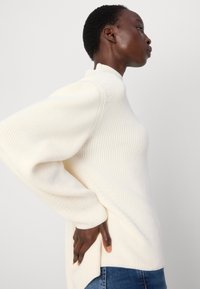 White ribbed knit sweater with a high collar and wide sleeves. Features side slits at the hem for ease of movement. Worn with blue jeans.