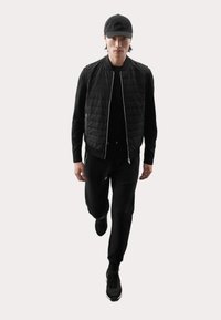 Zwarte gewatteerde vest over een zwart langärmige top, gecombineerd met zwarte broek en zwarte sportschoenen. Model draagt een zwarte pet.