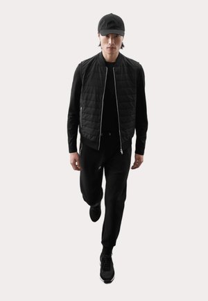 Black quilted vest over a black long-sleeve top, paired with black pants and black athletic shoes. Model wears a black cap.