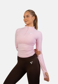 Różowa bluzka z długim rękawem z półzamkiem i otworami na kciuki, zestawiona z ciemnobrązowymi legginsami o wysokim stanie z akcentem w postaci logo.