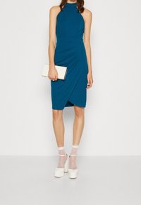 Türkisfarbenes ärmelloses Kleid mit hohem Kragen und drapiertem Design, kombiniert mit weißen Plateauschuhen und durchsichtigen Socken mit lila Punkten, während sie eine Clutch hält.