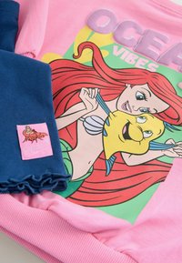 Rosa sweatshirt med tryck av "OCEAN VIBES" som visar en tecknad sjöjungfru, i kombination med blå volangleggings och en rosa etikett med en karaktärsillustration.