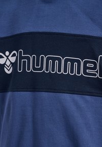 Tissu bleu avec une bande horizontale noire présentant le texte blanc contouré « hummel » et un logo stylisé d'abeille à gauche.