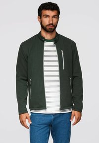 Grön texturerad jacka med hög krage, dragkedjefickor och en vit randig skjorta under. Blå denimbyxor fulländar looken.