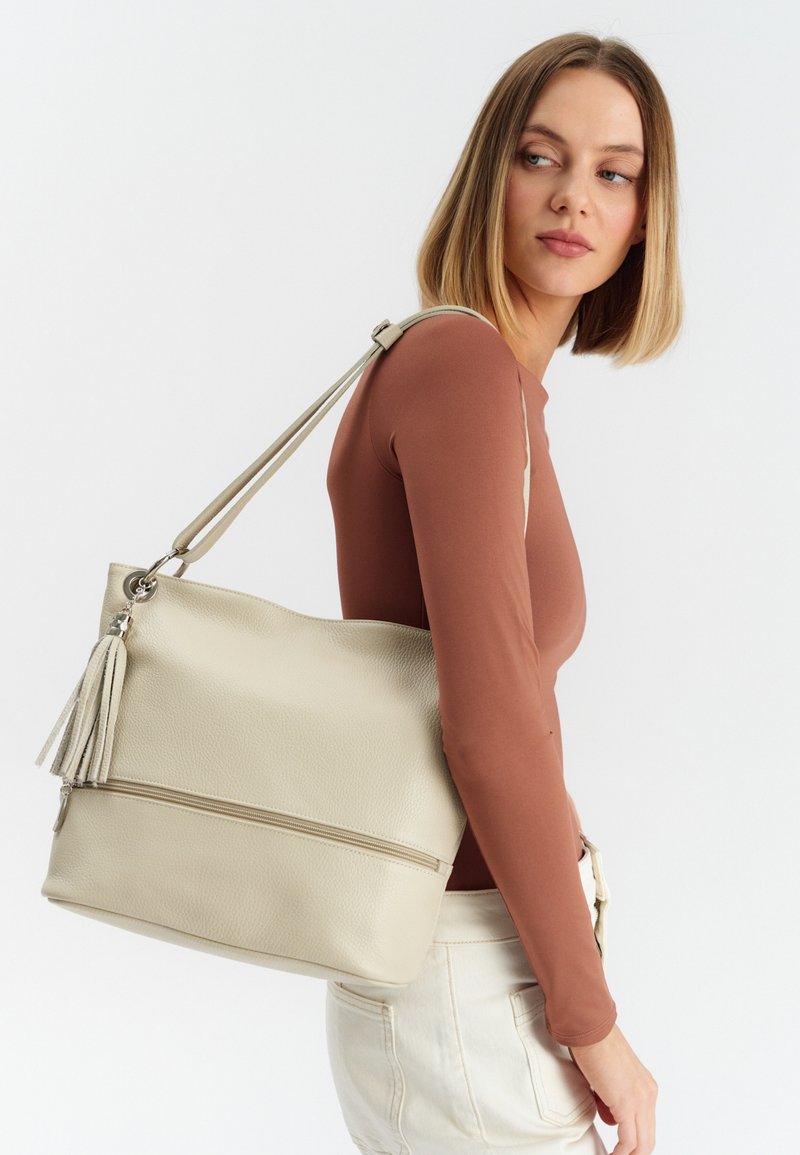 Sac fourre-tout en cuir beige clair avec une surface texturée, une seule bandoulière et un accent à pompon ; il possède une poche zippée à l'avant.