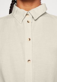 Camicia di lino beige chiaro con collo, dotata di cinque bottoni tondi e chiari lungo la parte anteriore e una superficie in tessuto testurizzato.