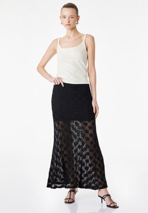 Femme portant un haut blanc sans manches et une longue jupe en dentelle noire avec des motifs floraux, associée à des sandales noires à talons.