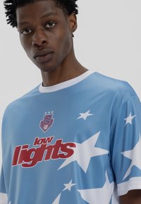 Ανοιχτό μπλε αθλητικό μπλουζάκι από λείο ύφασμα, με λευκά σχέδια αστέρων και κόκκινο κείμενο που αναγράφει "low lights". Λευκό γιακά και στολίδια στα μανίκια.