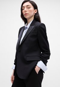 Zwarte op maat gemaakte blazer met satijnen revers, één knoop en zijzakken; gecombineerd met een lichtblauw gestreept overhemd, waarbij de manchet zichtbaar is.