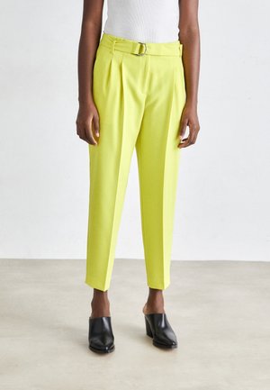 Persoon die hoog getailleerde, limoengroene geplooide broek met riem en zwarte mules met hak draagt, staand op een lichte vloer tegen een witte muur.