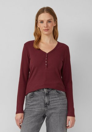 Burgunderrotes Langarmshirt aus geripptem Material mit V-Ausschnitt und einer Knopfleiste mit fünf Knöpfen, kombiniert mit hellgrauen Jeans im verwaschenen Design.