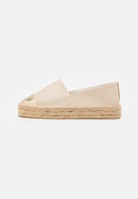 Beige espadrille dengan penutup jari kaki emas metallic dan sol jute anyaman. Bagian atas kulit halus dengan siluet datar dan desain slip-on.