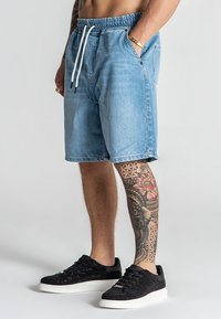 Shorts de mezclilla azul claro con cintura elástica y cordón; cuentan con bolsillos laterales y un ajuste relajado. Llevados con zapatillas negras de textura.