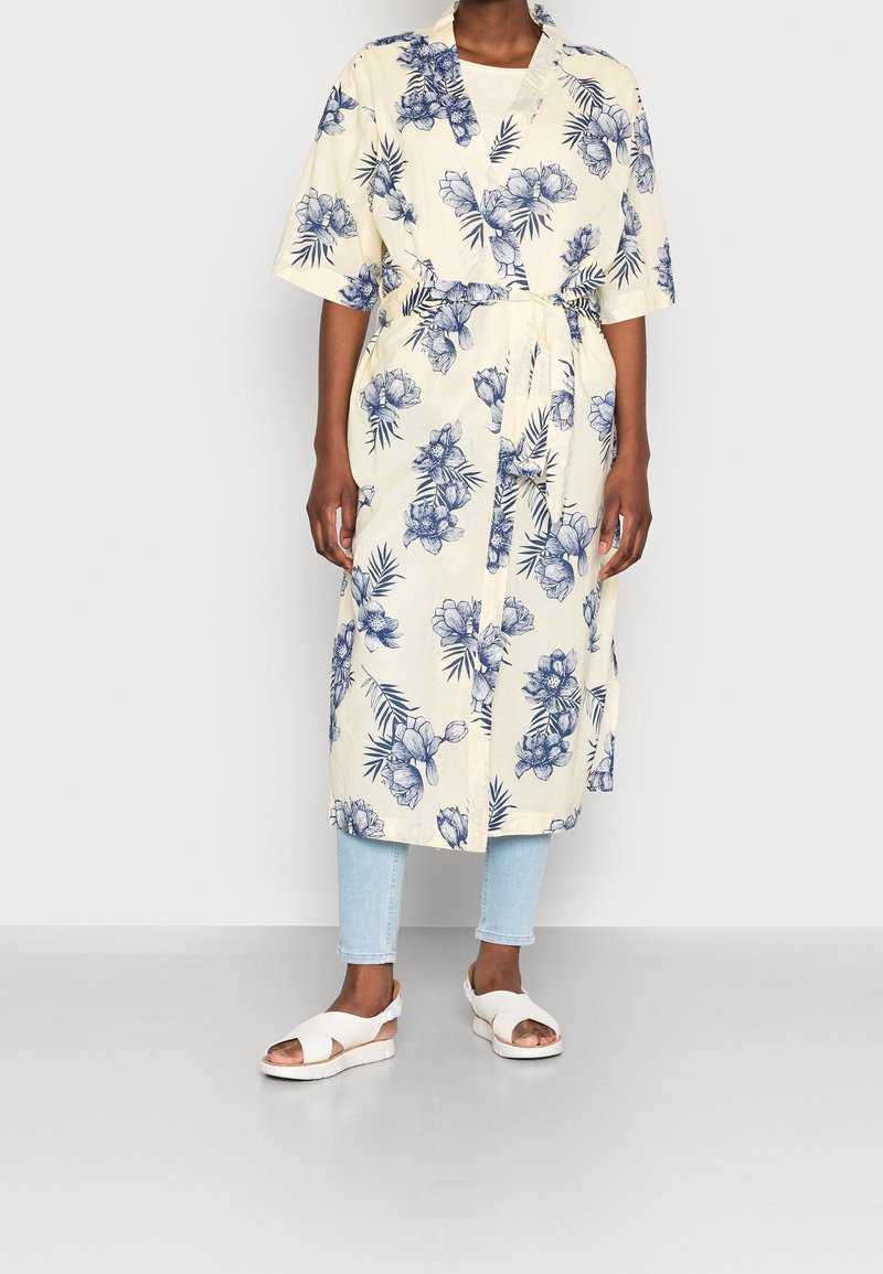 Kukkaisverhoiltu mekko vaaleassa kermanvärissä, jossa on sininen kasvitulostus, lyhyet hihat ja vyö. Yhdistetty vaaleansinisiin skinny-farkkuihin ja valkoisiin sandaaleihin.