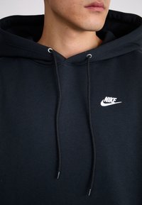 Black hooded sweatshirt made of cotton blend with a front pocket, white Nike logo on the left chest, and drawstring for hood adjustment.