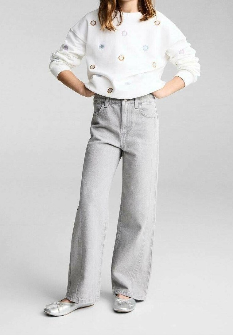 Fille debout, les mains sur les hanches, portant un pull blanc avec de petits motifs circulaires, un jean large gris clair et des ballerines argentées.