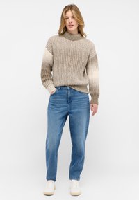 Suéter de punto beige con alto cuello y patrón texturizado, combinado con jeans de corte relajado azules y zapatillas de color crema.