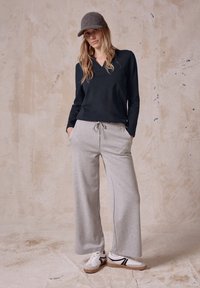 Svart långärmad bomullströja och grå vida sweatpants. Vita sneakers med svarta detaljer. Modellen står mot en strukturerad beige bakgrund.