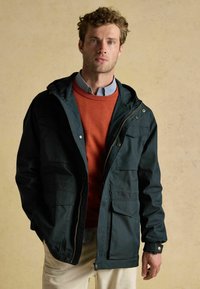 Jachetă cu glugă de culoare verde închis, cu fermoar și buzunare frontale, purtată peste un pulover portocaliu și o cămașă cu dungi, de către un bărbat pe un fundal neutru.