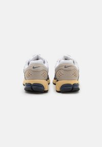 Nike-sneakers med en vitt meshuppsättning, beige häl med triangulära mönster och en gulaktig sula med blå accent.