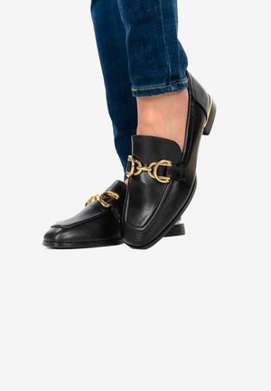 Personne portant des mocassins en cuir noir avec un détail mors doré et un jean bleu foncé sur un fond blanc.