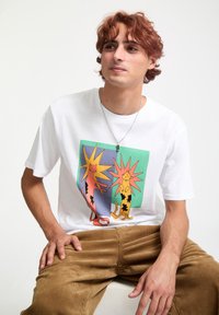 T-shirt blanc en coton avec un graphique coloré de deux chats stylisés entourés d'éclats de couleur vibrants. Associé à un pantalon en velours côtelé beige.