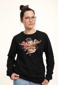 Černý svetr s dlouhými rukávy, který obsahuje barevně vyšitý design cherubínské postavy, obklopený textem a dekorativními prvky.