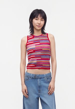 Jeune femme aux cheveux noirs portant un haut en tricot sans manches à rayures colorées et un jean bleu large, debout sur un fond blanc.
